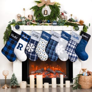 May include: Eight personalized Christmas stockings hanging in a row, featuring various blue, white, and grey patterns including buffalo plaid, snowflakes, and quilted textures. Each stocking is embroidered with a name: Violet, Rylee, Lilly, Marty, Andrew, River, Stella, and James.