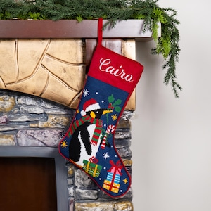 K&ouml;nnte beinhalten: Ein Weihnachtsstrumpf mit Pinguin-Design. Der Strumpf ist blau mit rotem Oberteil und zeigt einen Pinguin mit Weihnachtsm&uuml;tze und Schal, umgeben von Geschenken und Stechpalmen. Der Name "Cairo" ist oben eingestickt.