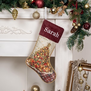 Op de afbeelding: Een kerstkous met een donkerrode ribfluwelen boord waarop de naam Sarah in het wit is geborduurd. Het lichaam toont een bedrukte kerstboom met ornamenten en cadeaus, afgewerkt met een rood geruite teen en gouden gevlochten randen.