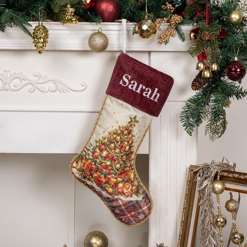 Op de afbeelding: Een kerstkous met een donkerrode ribfluwelen boord waarop de naam Sarah in het wit is geborduurd. Het lichaam toont een bedrukte kerstboom met ornamenten en cadeaus, afgewerkt met een rood geruite teen en gouden gevlochten randen.