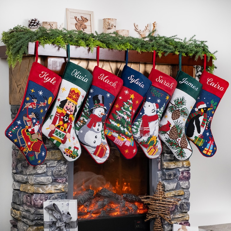 K&ouml;nnte beinhalten: Sieben personalisierte Weihnachtsstr&uuml;mpfe h&auml;ngen &uuml;ber einem Kamin. Jeder Strumpf hat ein anderes festliches Design und einen Namen auf der Manschette. Die Str&uuml;mpfe sind rot, gr&uuml;n und blau und zeigen verschiedene weihnachtliche Illustrationen.