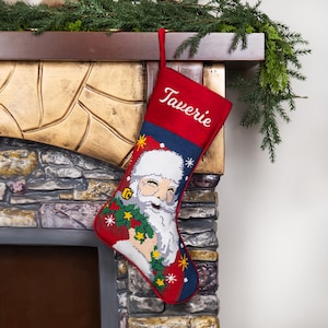 K&ouml;nnte beinhalten: Ein roter Weihnachtsstrumpf mit dem Namen "Taverie" in Wei&szlig; gestickt. Der Strumpf zeigt ein detailliertes Bild von Santa Claus, der einen Kranz h&auml;lt, mit einem blau-wei&szlig;en Hintergrund. Der Strumpf h&auml;ngt an einem Kamin.