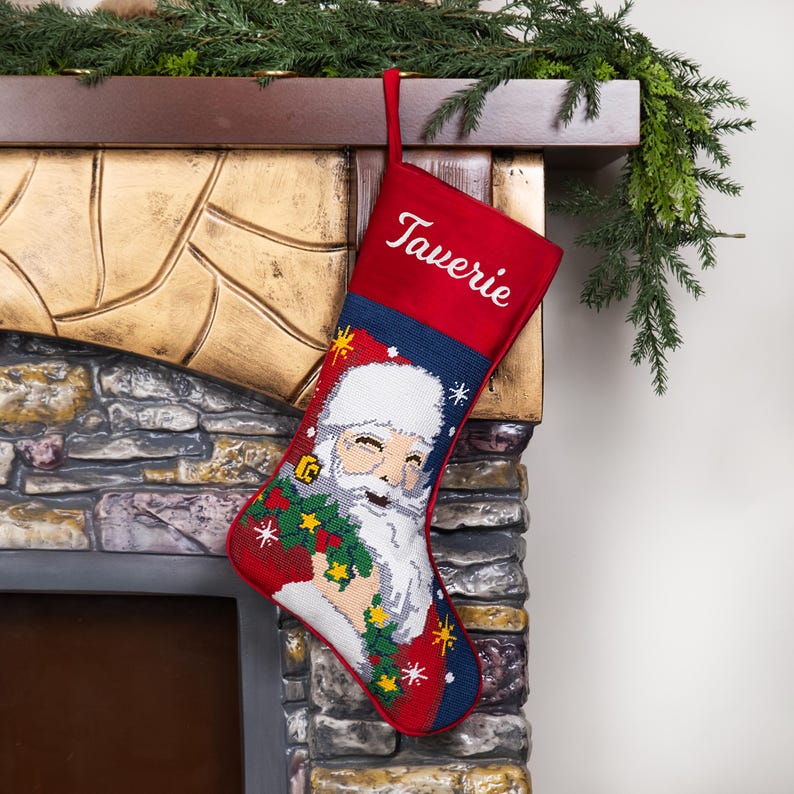 K&ouml;nnte beinhalten: Ein roter Weihnachtsstrumpf mit dem Namen "Taverie" in Wei&szlig; gestickt. Der Strumpf zeigt ein detailliertes Bild von Santa Claus, der einen Kranz h&auml;lt, mit einem blau-wei&szlig;en Hintergrund. Der Strumpf h&auml;ngt an einem Kamin.