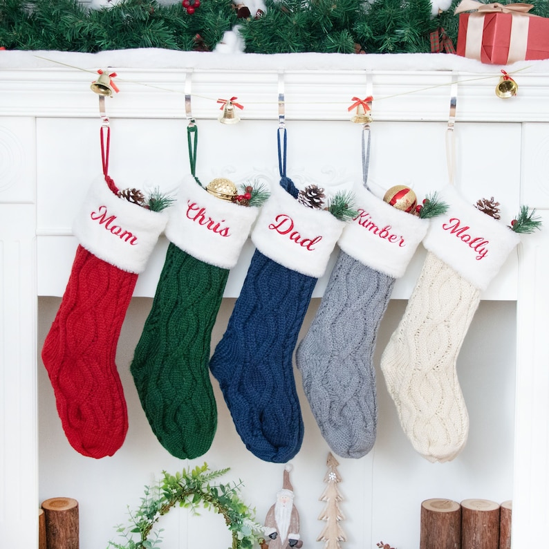 May include: Five knitted Christmas stockings with white faux fur trim hanging on a white mantel. The stockings are red, green, blue, gray, and white. Each stocking is personalized with a name: Mom, Chris, Dad, Kimber, and Molly.