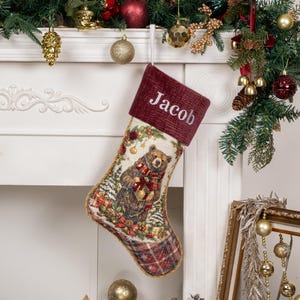 Op de afbeelding: Een kerstkous met een illustratie in tapijstijl van een bruine beer die een cadeau vasthoudt, met een donkerrode corduroy boord waarop de naam Jacob in het wit is geborduurd, afgewerkt met gouden gevlochten randen en een geruit teengedeelte.