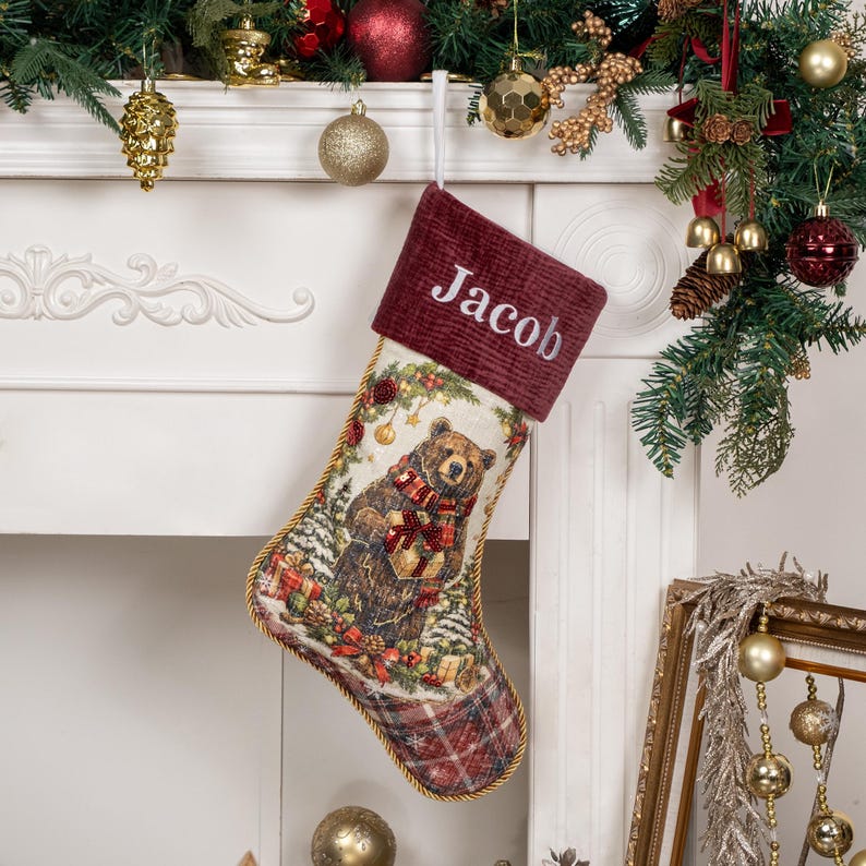 Op de afbeelding: Een kerstkous met een illustratie in tapijstijl van een bruine beer die een cadeau vasthoudt, met een donkerrode corduroy boord waarop de naam Jacob in het wit is geborduurd, afgewerkt met gouden gevlochten randen en een geruit teengedeelte.