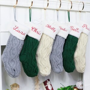 May include: Six personalized Christmas stockings with white faux fur trim and cable knit designs in gray, green, and cream. The stockings are hung on a white mantel and each has a name embroidered in red: Luna, Chris, Molly, Nimber, Bella, and Paw.