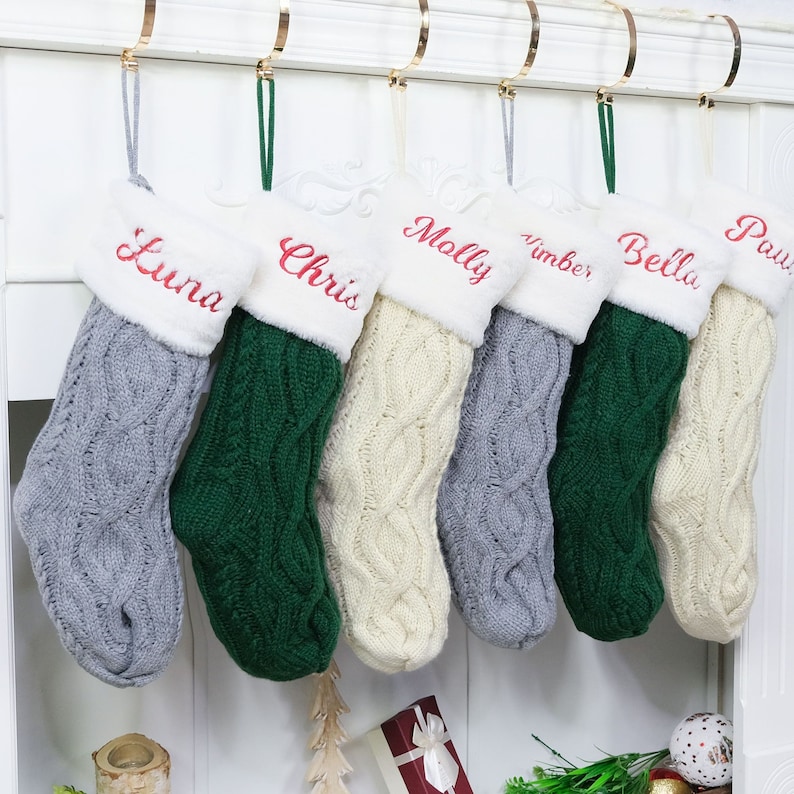 May include: Six personalized Christmas stockings with white faux fur trim and cable knit designs in gray, green, and cream. The stockings are hung on a white mantel and each has a name embroidered in red: Luna, Chris, Molly, Nimber, Bella, and Paw.