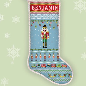 以下が含まれることがあります： 上部に赤い帯でBENJAMINという名前が入った、クリスマスストッキングのクロスステッチ図案。水色のストッキングには、くるみ割り人形、おもちゃの兵隊の列、カラフルな列車、ヒイラギのモチーフがデザインされています。
