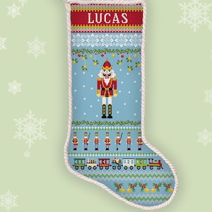 May include: A counted cross stitch Christmas stocking pattern featuring the name LUCAS in white text at the top. The design includes a central nutcracker, a row of toy soldiers, and a colorful train, all set against a light blue background.