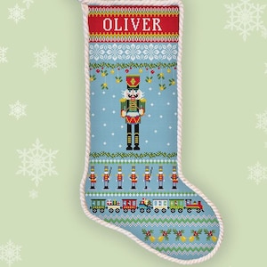 May include: A counted cross stitch pattern for a Christmas stocking featuring a Nutcracker and a train. The stocking is blue with red and white accents. The name "Oliver" is embroidered at the top of the stocking.