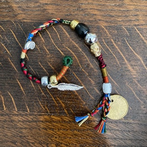 May include: A colorful braided bracelet with a skull charm, a feather charm, and a coin charm. The bracelet is made with a variety of colors, including red, green, blue, and yellow.