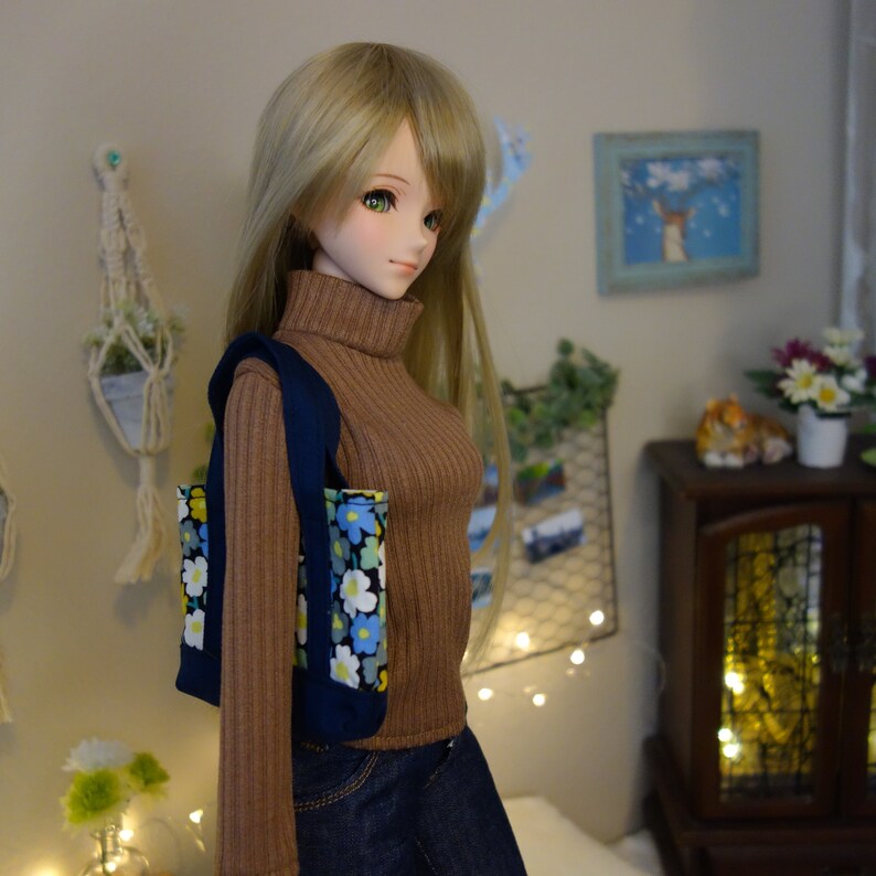 May include: A doll with long blonde hair wearing a brown turtleneck sweater and blue jeans. The doll is holding a blue and white floral tote bag.