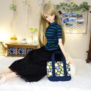 May include: A doll wearing a black skirt and a blue and green striped shirt, holding a small blue tote bag with a floral pattern.