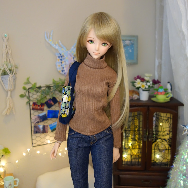 May include: A doll with long blonde hair wearing a brown turtleneck sweater and blue jeans. The doll is holding a floral print tote bag.