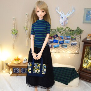 May include: A doll wearing a blue and green striped shirt, a black tulle skirt, and a floral tote bag. The doll is standing in front of a wall with a deer head decoration and a wire mesh wall with photos.