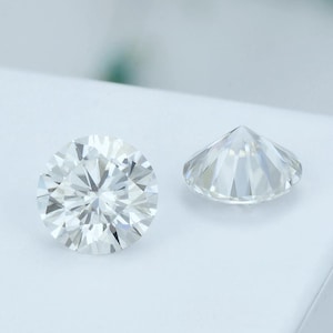 May include: Two round-cut, clear gemstones are displayed against a white background. The gemstones are faceted, with numerous triangular surfaces reflecting light. One stone is viewed from the top, showcasing its intricate facets, while the other is seen from the side.