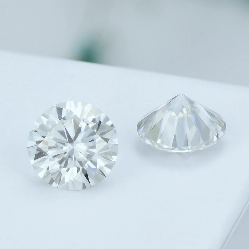 May include: Two round-cut, clear gemstones are displayed against a white background. The gemstones are faceted, with numerous triangular surfaces reflecting light. One stone is viewed from the top, showcasing its intricate facets, while the other is seen from the side.