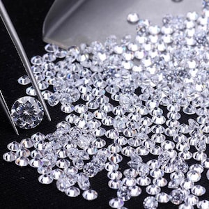 May include: A collection of small, clear, round gemstones, possibly diamonds, are scattered across a dark surface. A single, larger gemstone is held by a pair of tweezers. A small metal scoop is visible in the background, suggesting the gemstones are being sorted or handled.