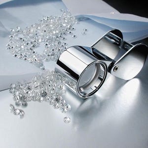 May include: A shiny, chrome-colored jeweler's loupe next to a scattering of small, clear gemstones. The loupe has two circular lenses and a hinged handle. The gemstones are scattered on a reflective surface.
