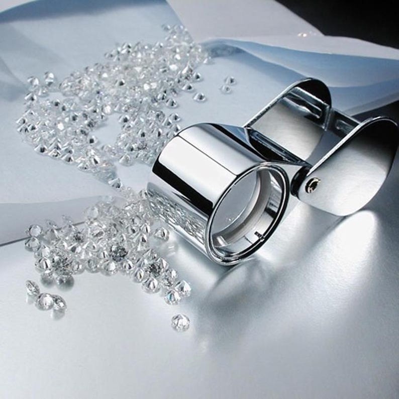 May include: A shiny, chrome-colored jeweler's loupe next to a scattering of small, clear gemstones. The loupe has two circular lenses and a hinged handle. The gemstones are scattered on a reflective surface.