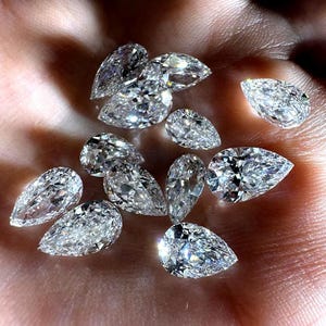 Peut inclure: Une collection de diamants en forme de poire, chacun avec de multiples facettes, est présentée. Les diamants sont clairs et réfléchissent la lumière, créant un effet scintillant. Les pierres précieuses sont tenues dans une main, avec le ton de la peau offrant un contraste chaleureux.