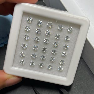 May include: A white plastic tray filled with numerous round, brilliant-cut diamonds. The diamonds are set against a light gray background, and the tray is held in a person's hand. The diamonds sparkle, reflecting light.