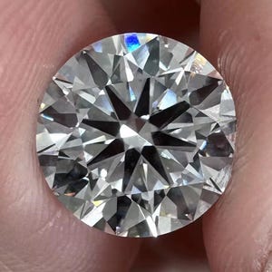 May include: A round, brilliant-cut diamond with numerous facets, reflecting light. The diamond is held between fingers, showcasing its clarity and sparkle. The gemstone's cut creates a star-like pattern in the center, highlighting its brilliance.