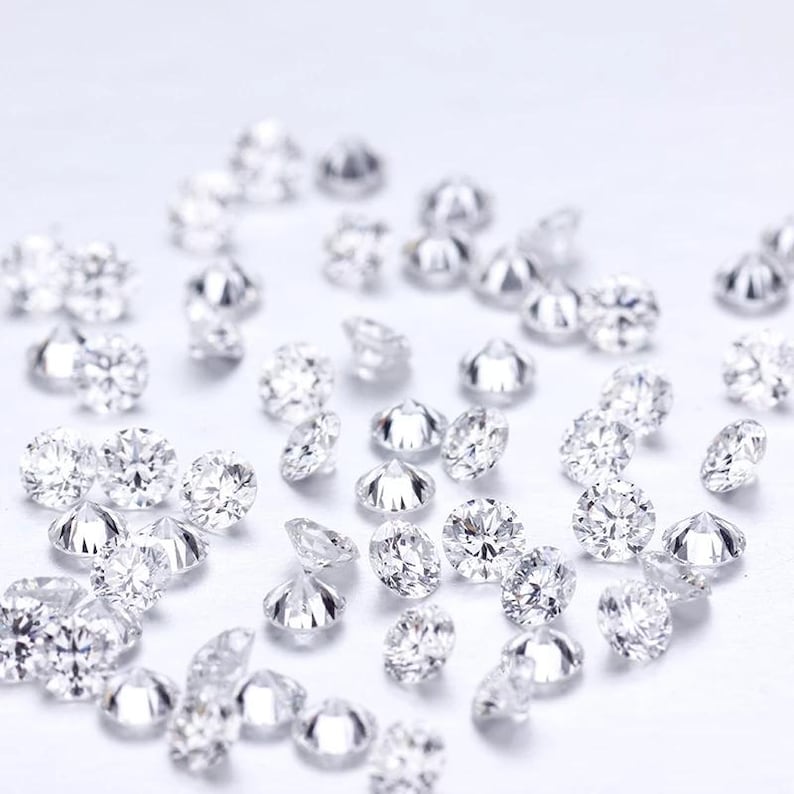 May include: A collection of sparkling, round-cut diamonds scattered across a white surface. The diamonds are clear and reflect light, showcasing their facets. The image highlights the brilliance and clarity of the gemstones, suggesting their use in jewelry or other decorative applications.