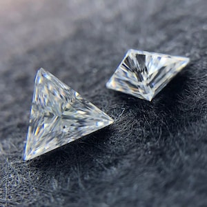 Pode incluir: Duas pedras preciosas transparentes e facetadas de forma triangular sobre uma superfície texturizada cinza escura. A maior pedra preciosa é um triângulo, e a menor é um quadrado.