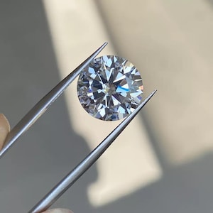 May include: A round, brilliant-cut diamond held by silver tweezers. The diamond sparkles with numerous facets, reflecting light. The tweezers are positioned at an angle, showcasing the gem's clarity and brilliance. The background is a soft, neutral tone, highlighting the diamond's sparkle.