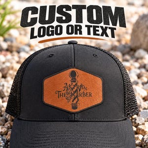 May include: A black trucker hat featuring a hexagonal tan leather patch on the front. The patch displays a graphic of a barber pole and the text "Astrid The Barber" in black. The hat has a solid front panel and a mesh back with a curved brim.
