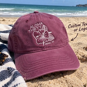 May include: A maroon baseball cap featuring white embroidered text that reads ALOHA THIRTY and MAUI, HI, accompanied by a white hibiscus flower graphic. The cap has a curved brim and a button on top, with a visible arrow pointing to the cap.
