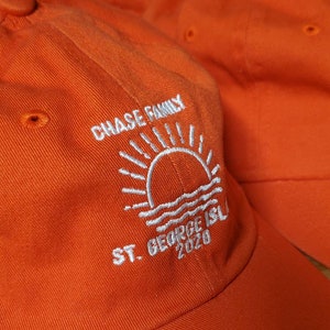 May include: Bright orange baseball caps with white embroidered text and a sun and water design. The text reads "CHASE FAMILY", "ST. GEORGE ISL" and "2020". The caps are made of a durable fabric.