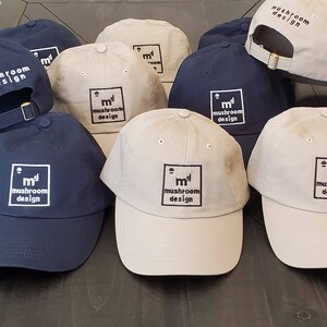 May include: A collection of baseball caps in various colors, including navy blue and beige. Each cap features the text "mushroom design" within a square frame. The caps have a curved brim and an adjustable strap.