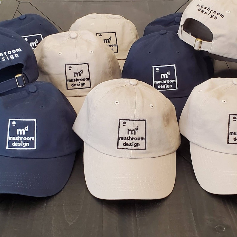 May include: A collection of baseball caps in various colors, including navy blue and beige. Each cap features the text "mushroom design" within a square frame. The caps have a curved brim and an adjustable strap.