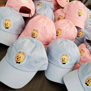 May include: A collection of baseball caps in light blue and pink. Each cap features an embroidered design of a dog wearing a bow tie. Some caps have the text "Pete's Pints" embroidered on the back.