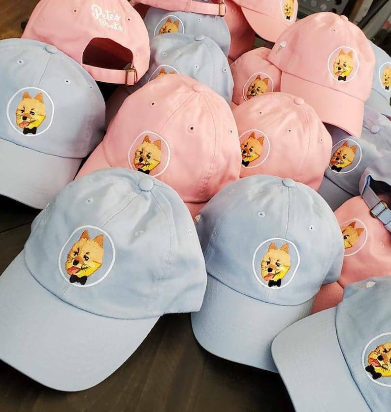 May include: A collection of baseball caps in light blue and pink. Each cap features an embroidered design of a dog wearing a bow tie. Some caps have the text "Pete's Pints" embroidered on the back.