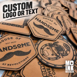 May include: A collection of tan leather patches featuring custom black laser-engraved designs, including text such as HOUSE OF HANDSOME EST 2019 AT REVOLUTIONS, a mustache graphic, and a vintage diving helmet illustration, with a MO INK CUSTOM logo.