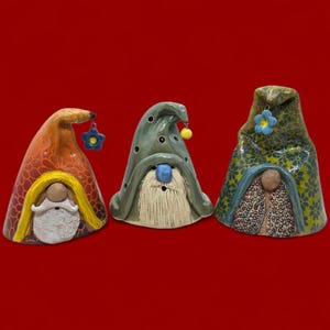 May include: Three ceramic gnome figurines with pointed hats and long beards. The gnomes have different hat designs and colors, including orange, green, and blue. Each gnome has a flower or a yellow ball hanging from its hat.