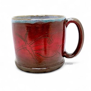 May include: A handmade ceramic mug with a deep red glaze and a dark brown base and handle. The mug features an etched design of a butterfly or moth. The rim has a blue-green glaze. The text "LMSR1" is on the side of the mug.