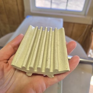 May include: A speckled, light yellow ceramic soap dish with vertical ridges and small feet. The dish is held in a hand, showcasing its curved shape and textured surface. The design is simple and functional.
