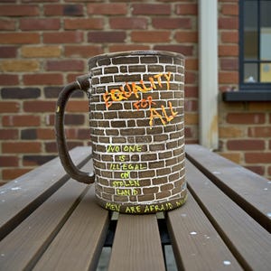 May include: A brown ceramic mug with a brick wall design and the words "EQUALITY FOR ALL", "NO ONE IS ILLEGAL ON STOLEN LAND", and "MEN ARE AFRAID NOW" in orange and yellow lettering. The mug has a brown handle and sits on a wooden table.