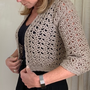 May include: A beige crocheted cardigan with a delicate openwork pattern. The cardigan has a V-neck and three-quarter sleeves.