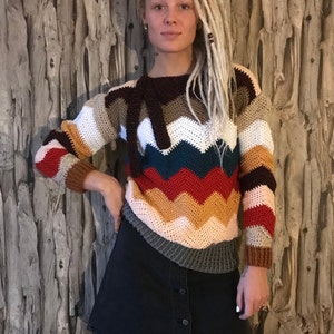 May include: A woman wearing a multi-coloured crocheted sweater with a chevron pattern. The sweater has stripes of brown, white, teal, red, orange, pink, and grey. She is also wearing a black denim skirt.
