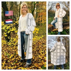 Könnte beinhalten: Ein langer, weißer Häkelcardigan mit grauen Streifen. Der Cardigan wird von einer Person getragen, die in einer Waldlandschaft steht. Der Cardigan hat einen V-Ausschnitt und lange Ärmel.