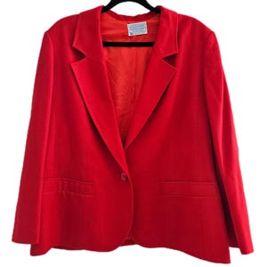 Könnte beinhalten: Ein leuchtend roter Blazer mit einem Ein-Knopf-Verschluss und zwei Vordertaschen. Der Blazer hat ein klassisches Reversdesign und ein rotes Innenfutter. Ein Etikett ist in der Nähe des Kragens sichtbar. Der Blazer hängt an einem schwarzen Kleiderbügel.