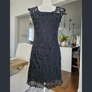 May include: A black sleeveless dress featuring a sheer overlay with a tonal embroidered floral and vine pattern. The garment has a scoop neckline, a fitted bodice, and a straight hemline that falls above the knee, showcasing a solid black lining.