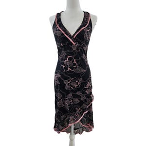 Puede incluir: Vestido negro sin mangas con escote en V y estampado floral rosa. El vestido tiene un dobladillo con volantes y ribete rosa. El vestido se exhibe en un maniquí. El vestido mide aproximadamente 100 cm de largo.