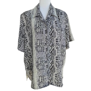 May include: A short-sleeved button-down shirt featuring a monochromatic snakeskin print in shades of grey, black, and white. The garment has a classic collar, a front button closure, and a repeating reptilian scale pattern across the fabric.